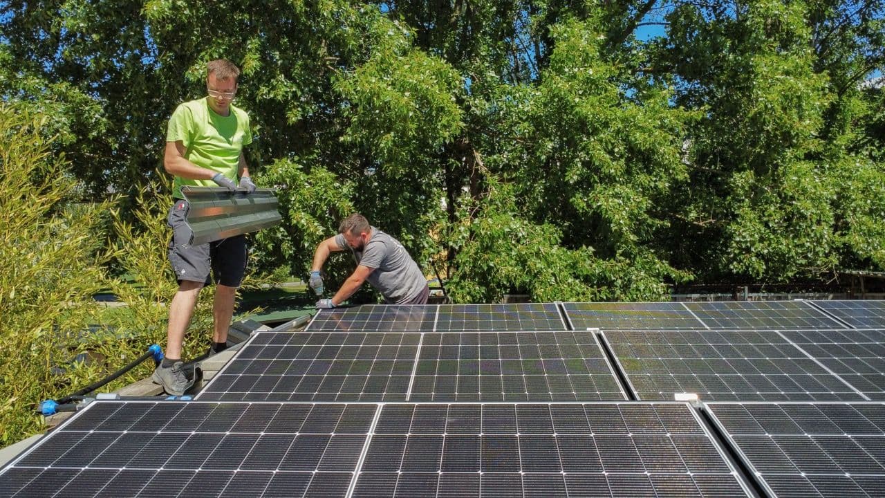 Ein Mitarbeiter der Firma sunday solar trägt einen Träger für eine Solaranlage, ein weiterer fixiert gerade in Solarpanel. Drei Reihen an Photovoltaik Modulen sind zu sehen. Dahinter grüne Bäume.